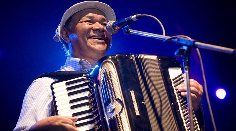 O músico Dominguinhos sorridente no palco tocando sua sanfona preta diante do microfone, ilustrando uma música