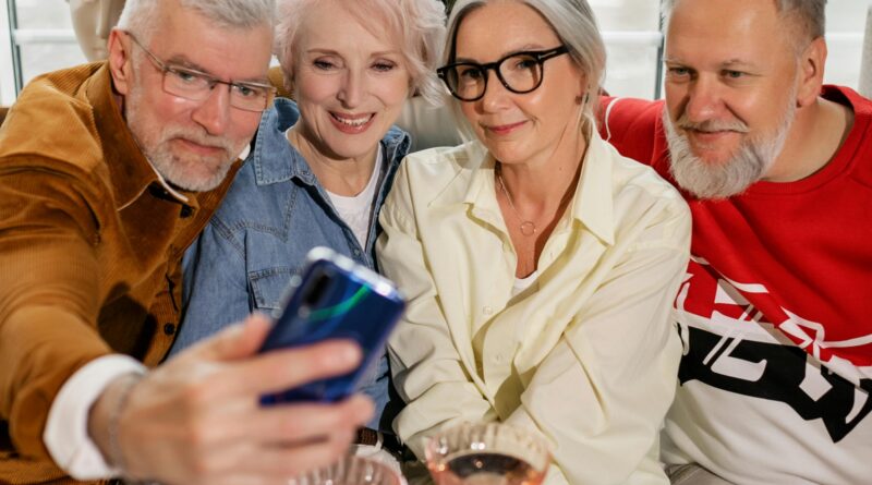 Dois casais de idosos sorridentes reunidos ao redor de uma mesa, tirando uma selfie com o celular para celebrar a amizade na terceira idade.
