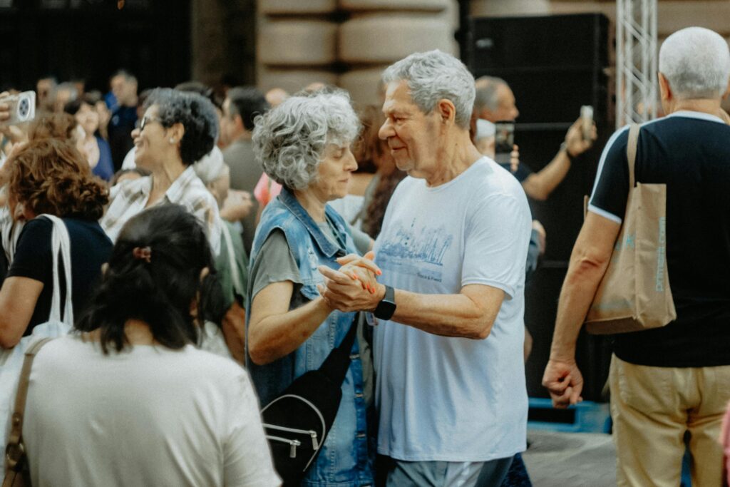 Casal de idosos felizes dançando sorridentes em um salão durante um animado baile da terceira idade com casais ao fundo.
