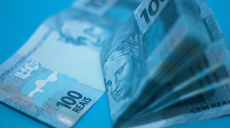 Duas cédulas de cem reais azuis do Brasil sobre uma mesa, sendo uma esticada e um maço dobrado, representando os valores a receber esquecidos no banco.