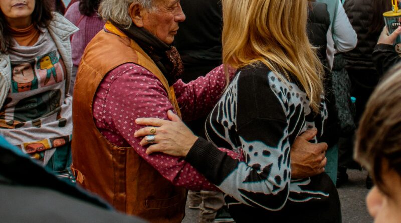 Casal de idosos felizes dançando sorridentes durante um animado baile da terceira idade com casais ao fundo.