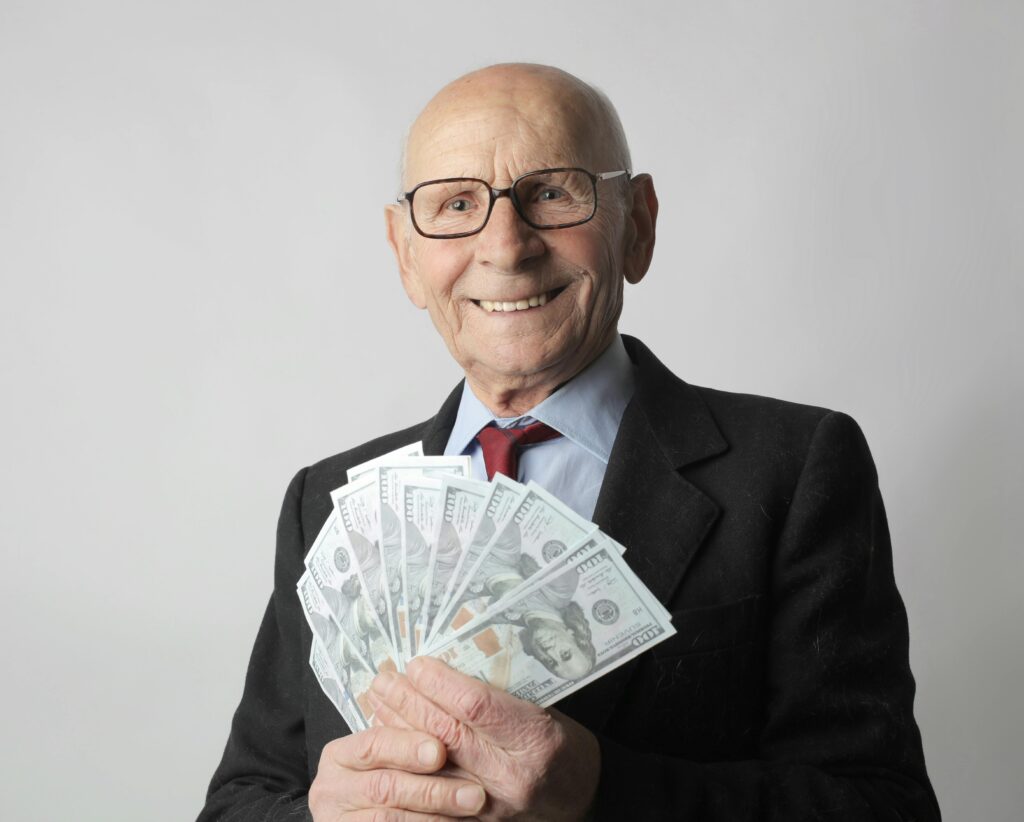 Homem idoso sorridente vestindo terno preto e óculos, segurando várias notas de dinheiro em formato de leque, ilustrando o resgate de valores a receber.