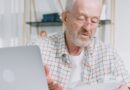 Homem idoso de óculos olhando confuso para a tela de um computador portátil na mesa de casa, analisando o extrato do seu benefício previdenciário.